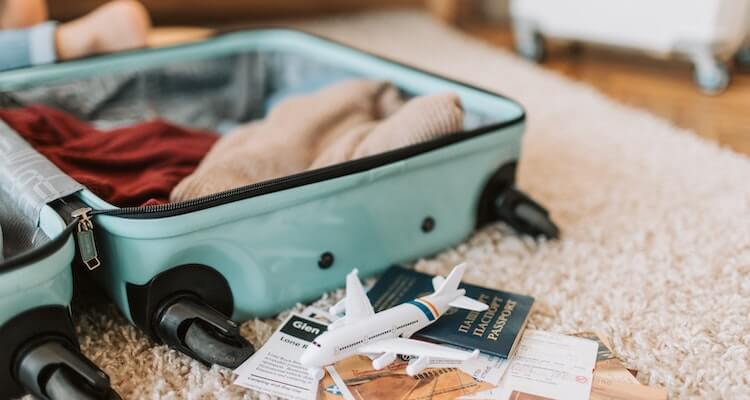 A suitcase open on the floor mid-packing. You can see sweaters inside the suitcase and a passport and model airplane next to it.